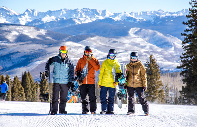 A group of four snowboarders stands at the top of a run holding their boards
