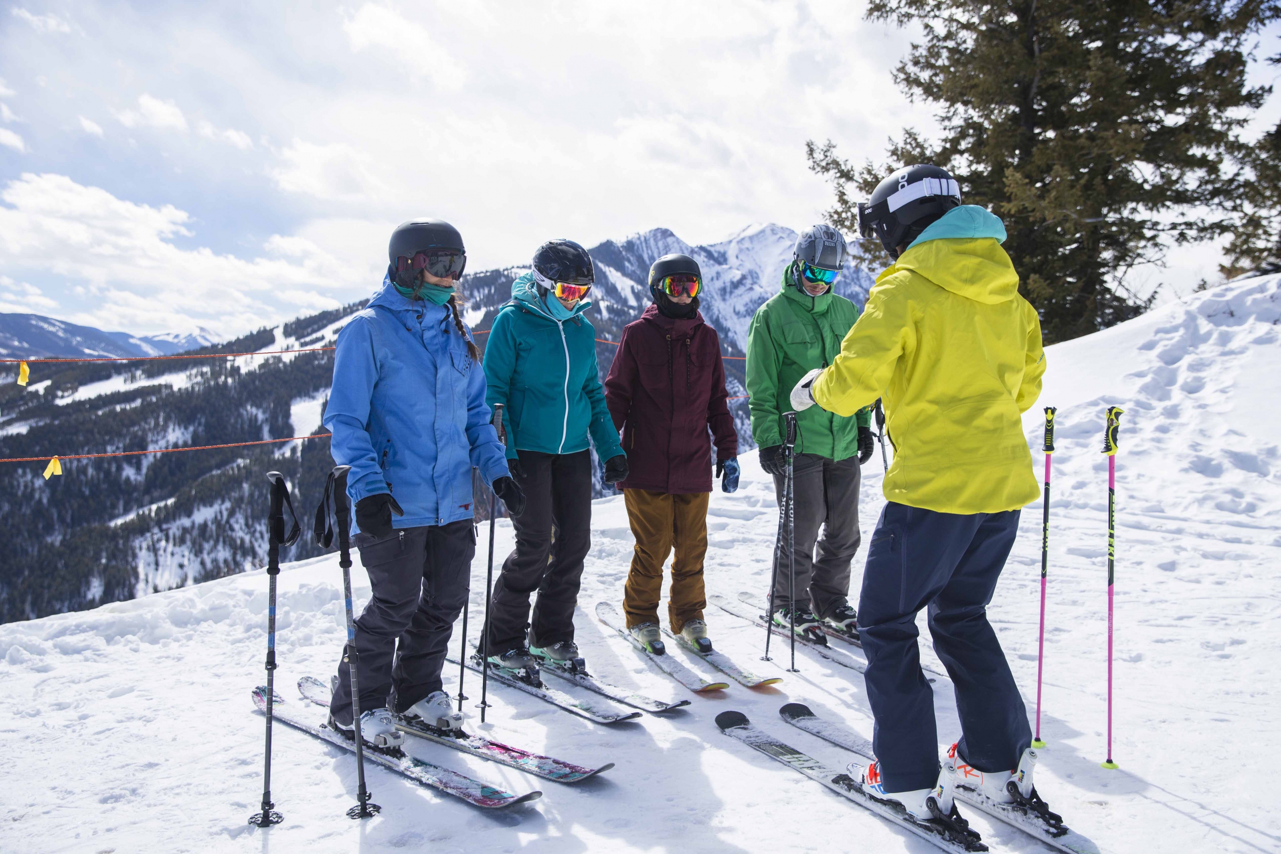An alpine instructor talks to their four students at the top of a run