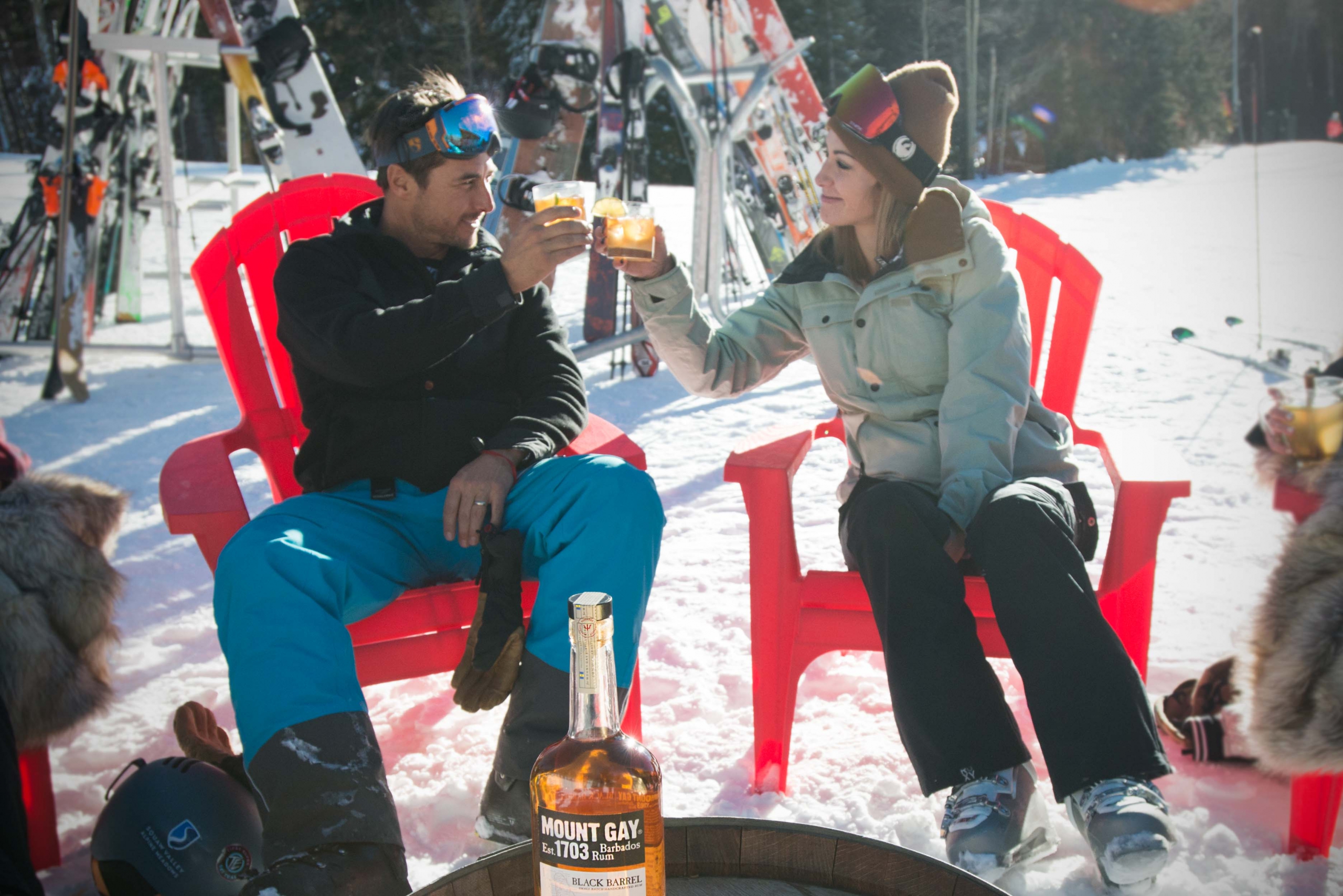 A man a women sit in red chairs at the base of a ski slope as they cheers their cocktails