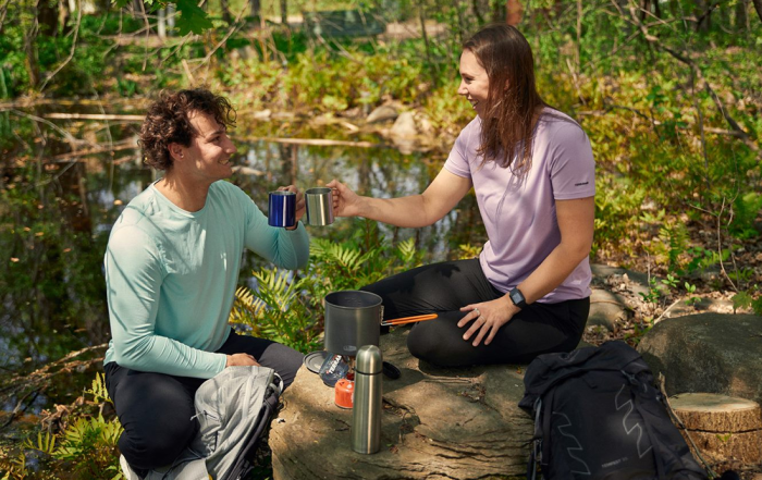 A man and women drink a hot beverage heated by a camping stove while sitting in front of a lake.