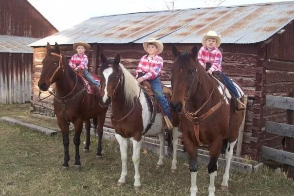 Three boys dressed in western wear on horses in front of a barn.