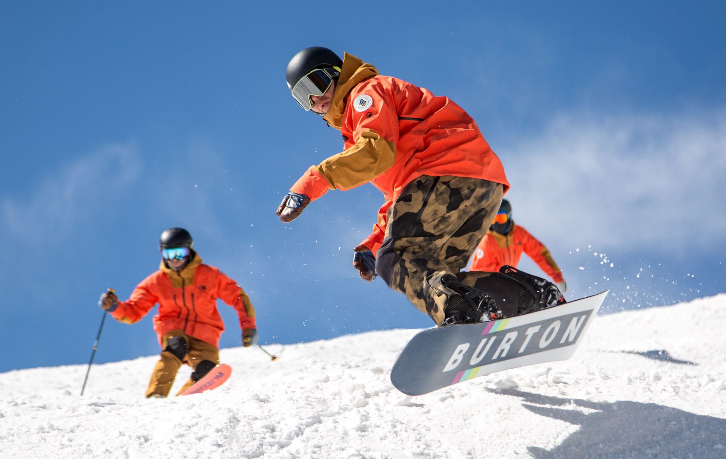 Snowboarder jumping in red jacket with skier in the background.